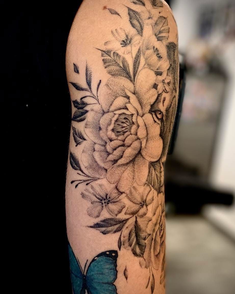 Tatouage floral noir et gris sur le haut du bras, représentant des pivoines et des feuilles détaillées au-dessus d'un papillon bleu.