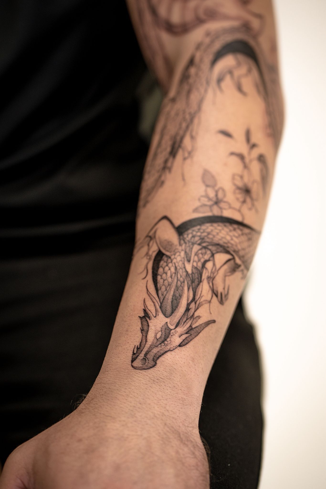 L'avant-bras d'une personne orné d'un tatouage détaillé à l'encre noire représentant une créature ressemblant à un dragon et de délicats motifs floraux.