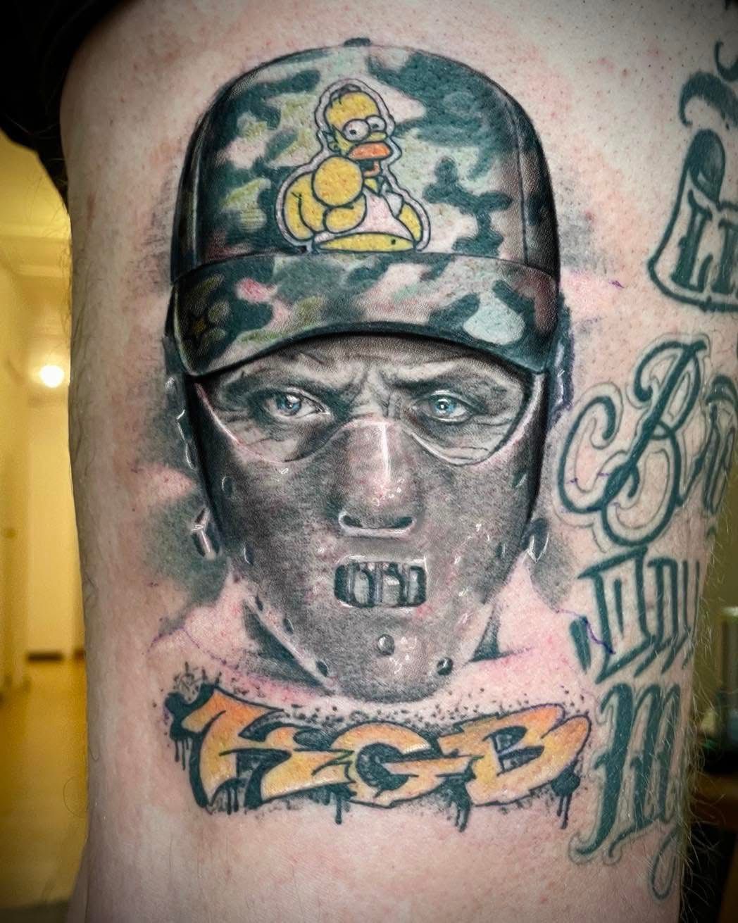 Un tatouage représentant une personne portant un masque facial à la Hannibal Lecter et une casquette camouflage ornée d'un dessin d'Homer Simpson.