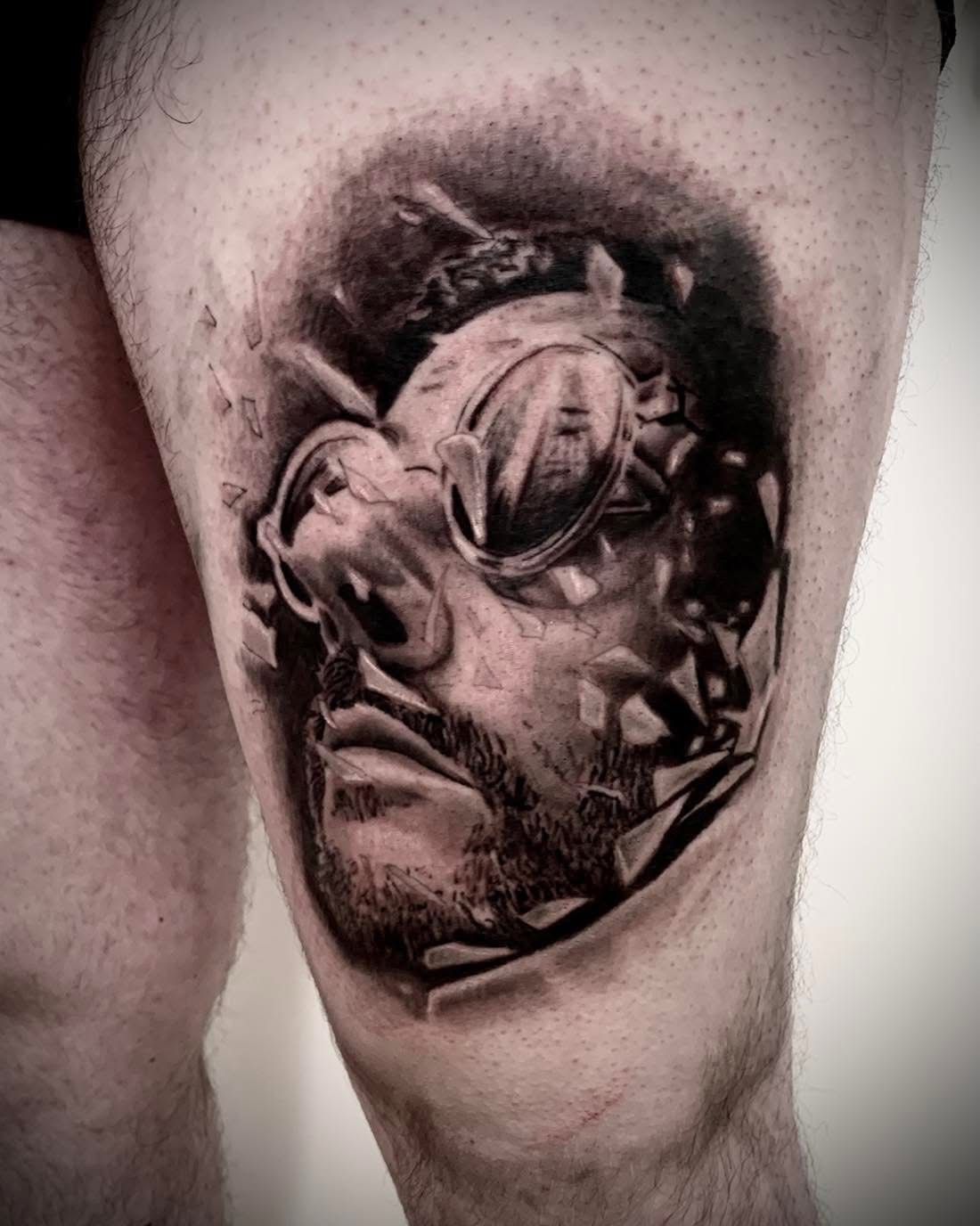 Un tatouage en noir et gris sur une cuisse représentant une personne portant des lunettes rondes, entourée d'éclats de verre flottants.