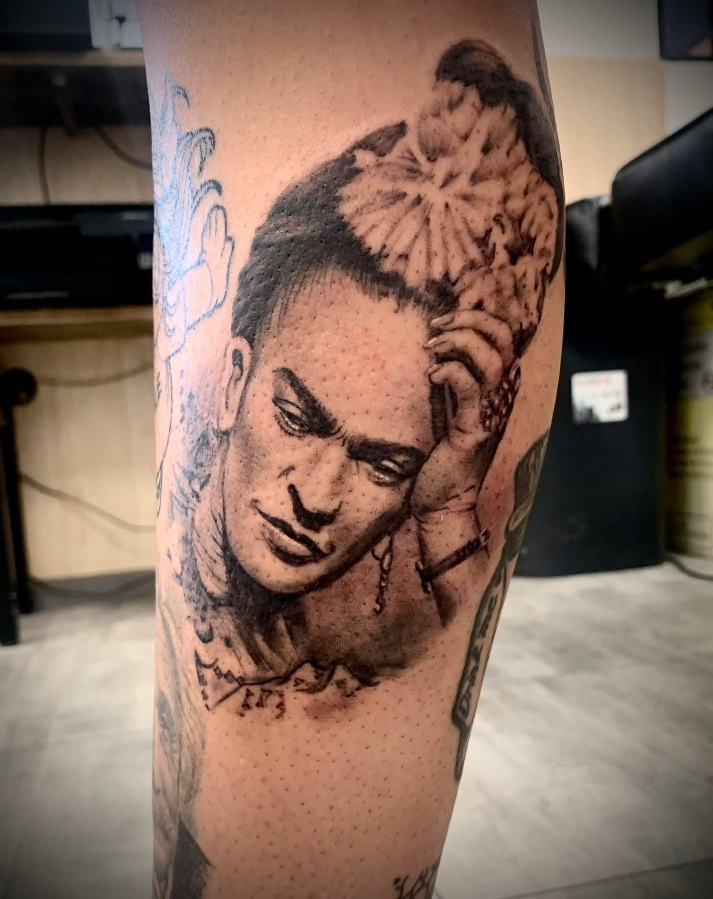 Un tatouage en noir et gris représentant Frida Kahlo, l'air pensif, la tête appuyée sur sa main ornée de motifs floraux.
