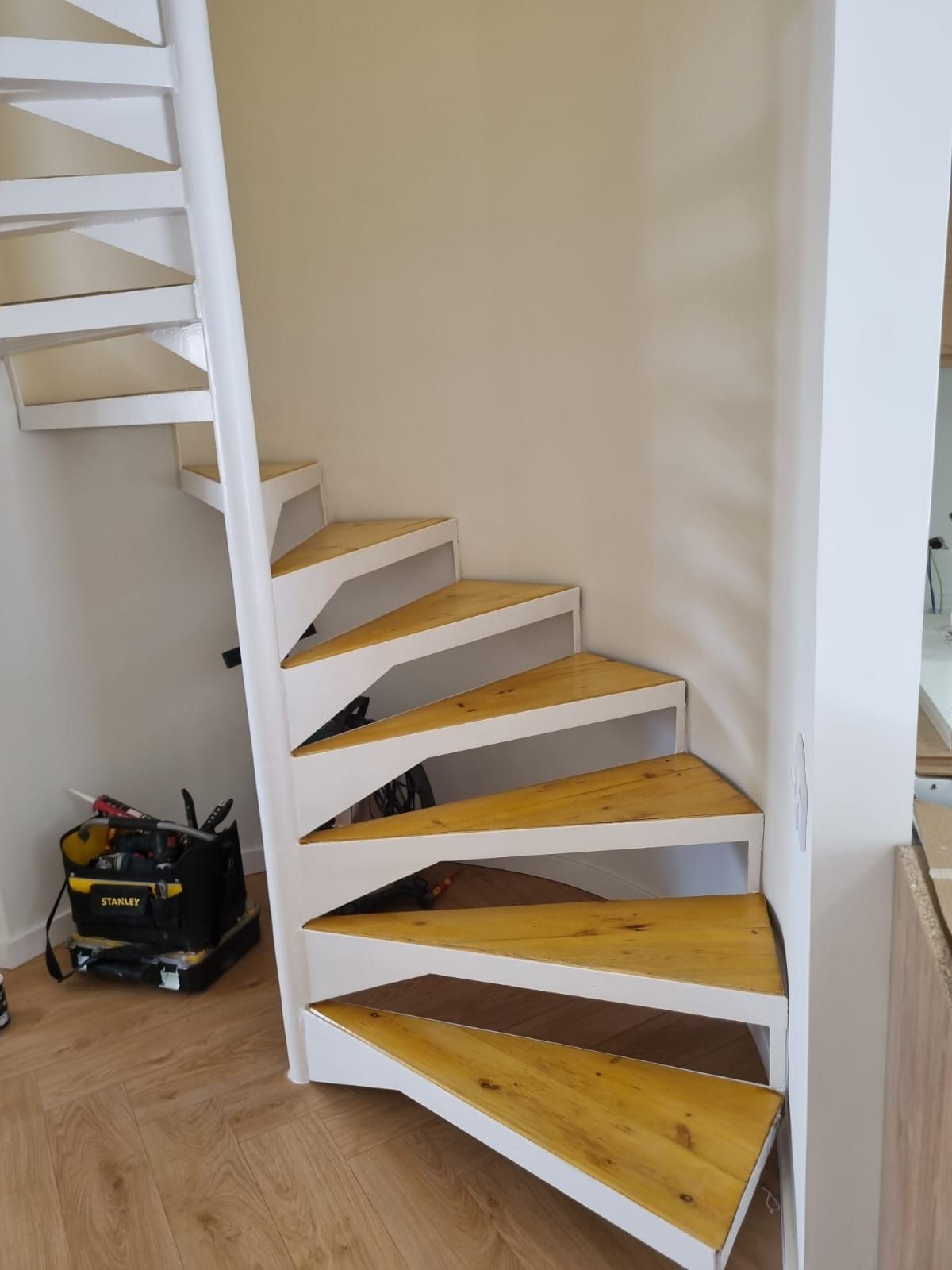 Escalera de caracol con marco blanco y peldaños de madera, contra una pared beige. Hay herramientas cerca.
