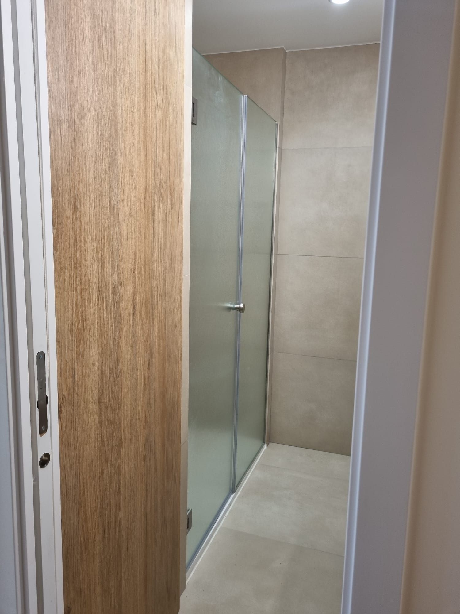 Interior de baño con puerta de madera, ducha de vidrio esmerilado y azulejos de tonos neutros.