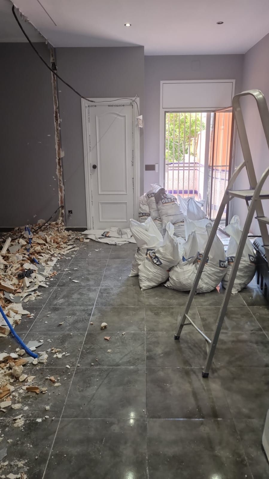 Habitación en remodelación, escombros en el piso, bolsas blancas, paredes grises, puerta, ventana, escalera.