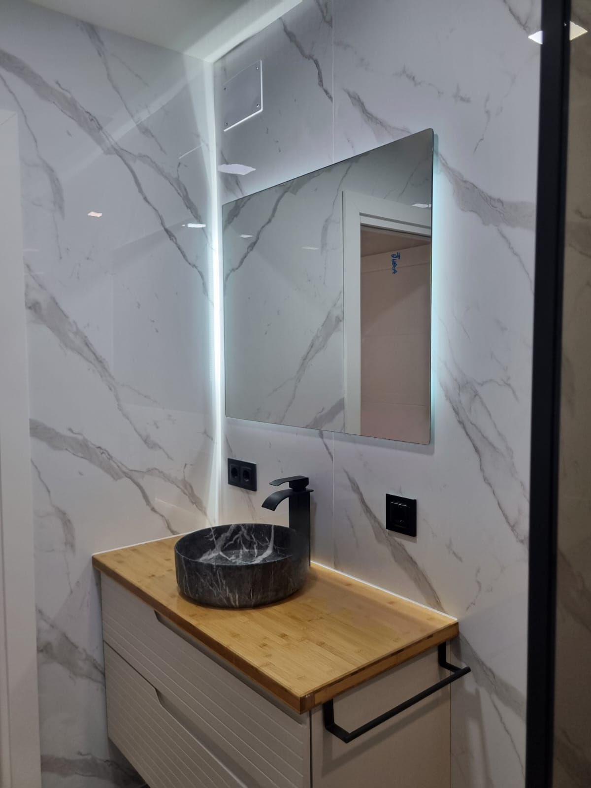 Baño moderno con paredes efecto mármol, tocador de madera con lavabo negro, espejo con luces y grifería negra.