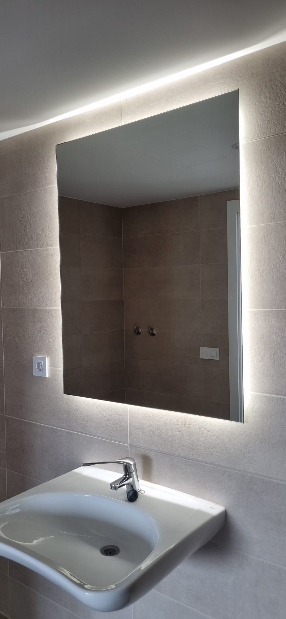 Espejo de baño con iluminación integrada sobre un lavabo blanco. Paredes de azulejos beige.