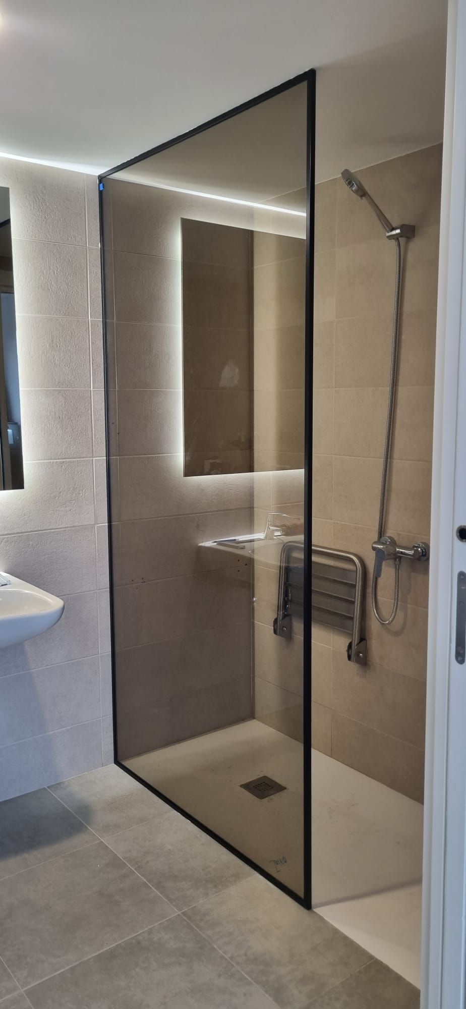 Baño moderno con mampara de ducha de cristal, azulejos grises y accesorios blancos.