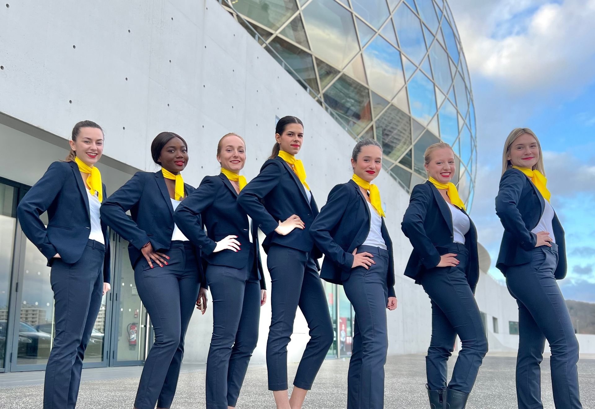Sept hôtesses vêtues de bleu au foulard jaune posent devant un grand bâtiment