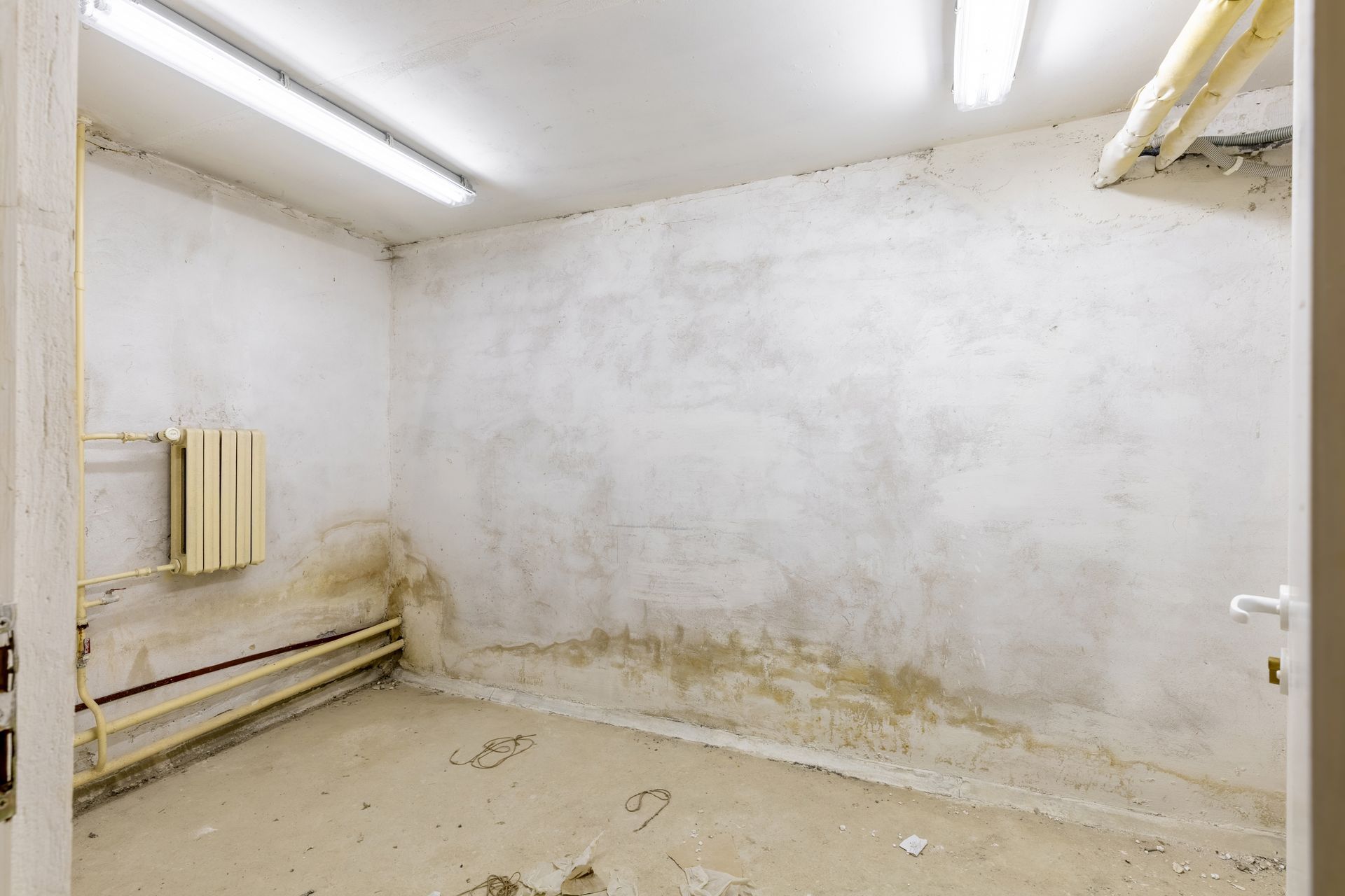 Une petite pièce humide aux murs blancs écaillés, avec des tuyaux apparents et un radiateur, évoquant une cave