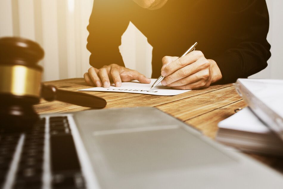 Persona escribiendo en papel con un bolígrafo en un escritorio de madera con un mazo, un ordenador portátil y libros.