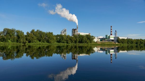 Baustoffindustrie Industrieanlage mit Schornsteinen an einem Fluss, die sich im ruhigen Wasser unter einem klaren blauen Himmel spiegeln.