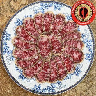 Salchichón ibérico curado loncheado sobre plato floral azul y blanco, con logo rojo.