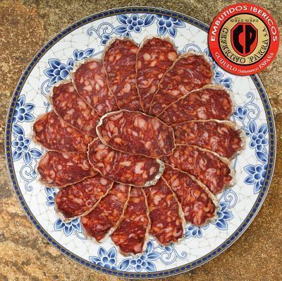 Rebanadas de chorizo español dispuestas en un plato con estampado azul y blanco; etiqueta del producto en la esquina.