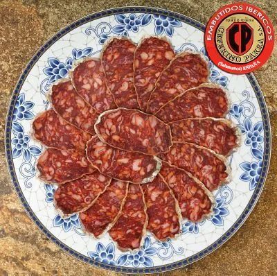 Rebanadas de chorizo español dispuestas en un plato con estampado azul y blanco; etiqueta del producto en la esquina.