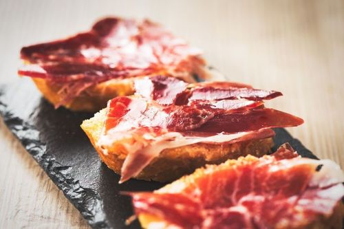 Tostada cubierta con finas lonchas de jamón curado, dispuestas sobre una tabla de pizarra negra.