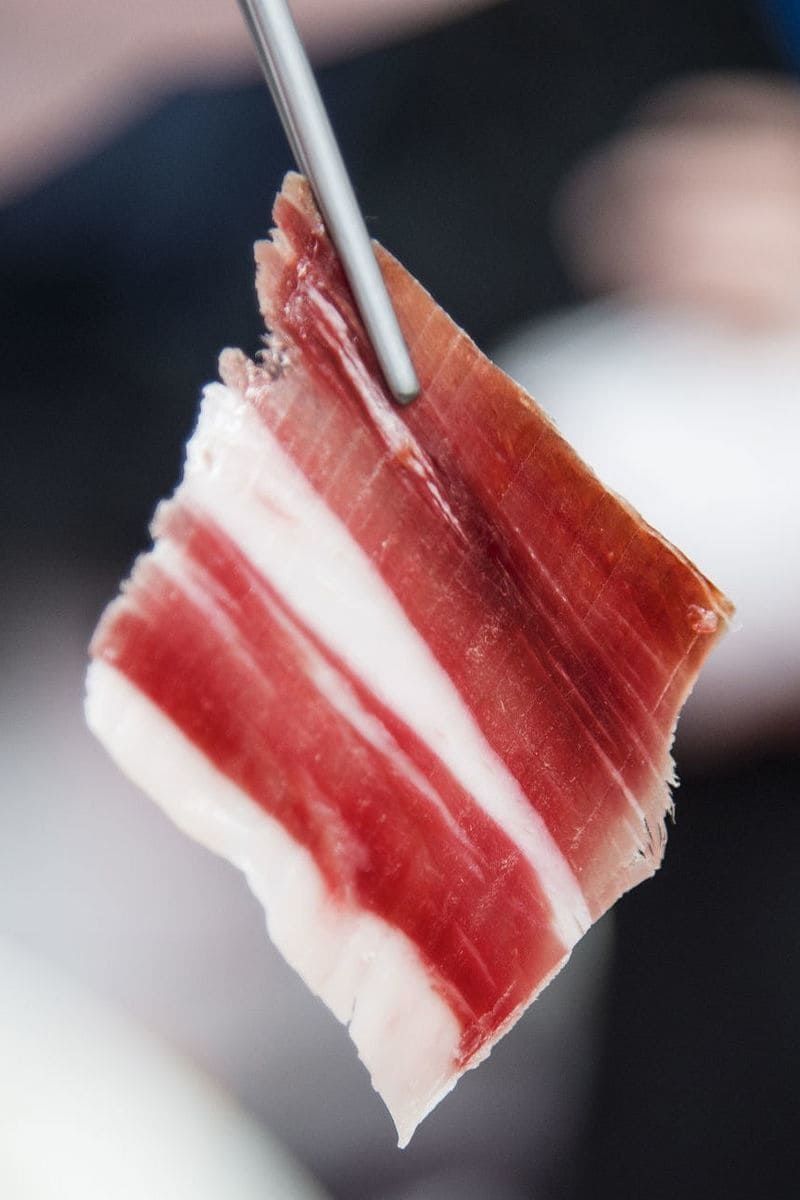 Loncha fina de jamón ibérico, sujeta mediante pinzas metálicas, mostrando vetas rojas y blancas.