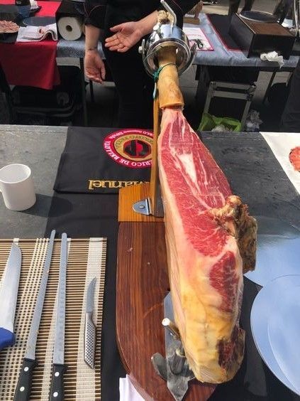 Una pata de jamón curado sobre un soporte de madera, lista para ser cortada, con cuchillos, una persona y una mesa.