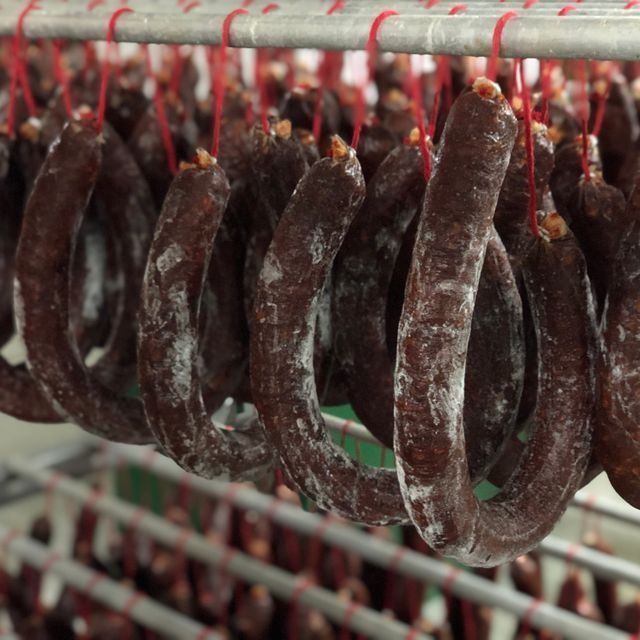Salchichas colgantes: eslabones curados de color marrón oscuro con moho blanco, suspendidos de un hilo rojo sobre soportes metálicos.