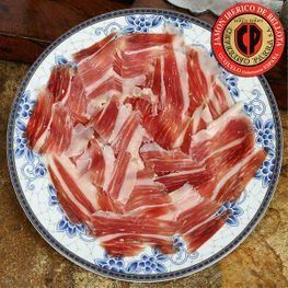 Jamón ibérico de bellota cortado en lonchas finas sobre plato estampado, con etiqueta roja y dorada.