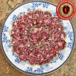 Salchichón ibérico curado loncheado sobre plato floral azul y blanco, con logo rojo.