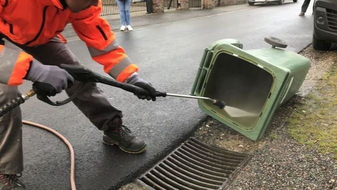 Un ouvrier en tenue de sécurité orange arrose au tuyau une poubelle verte renversée près d’une bouche d’égout.