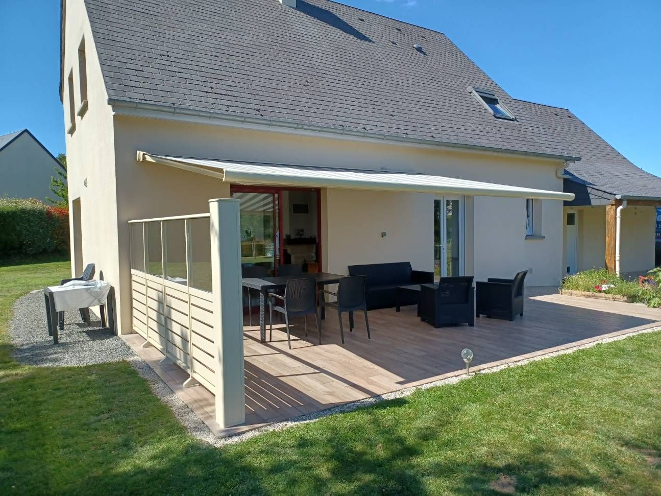 Une maison moderne avec terrasse, salon de jardin et auvent rétractable. Pelouse verte et ciel ensoleillé.