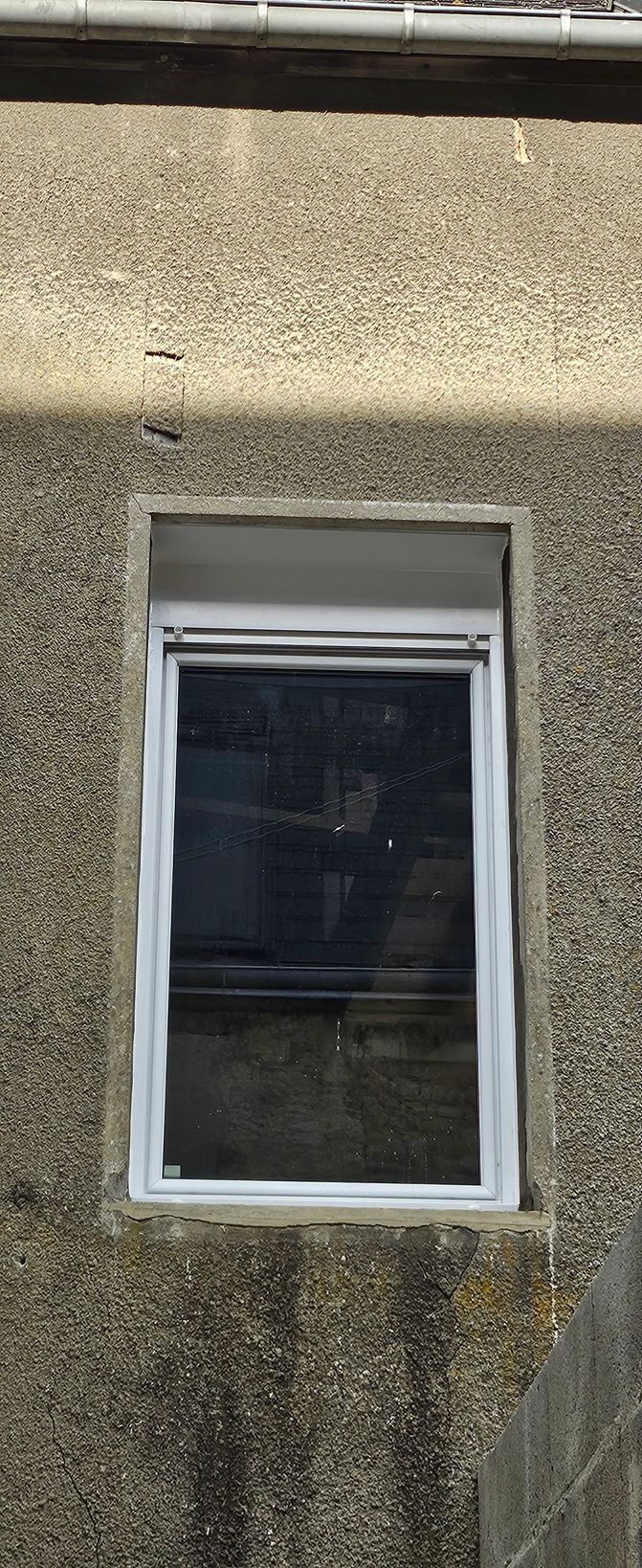 Fenêtre sur un bâtiment texturé et de couleur claire, avec un store foncé partiellement enroulé et un cadre blanc.