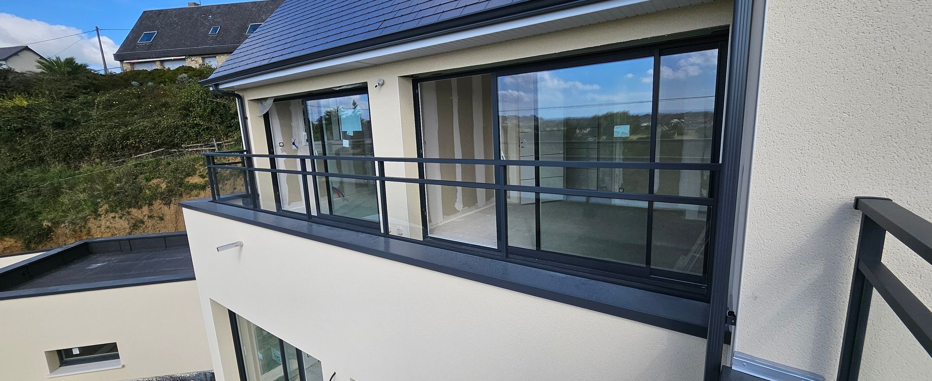 Vue extérieure d'une maison moderne avec balcon, dotée de grandes fenêtres et d'une balustrade noire.