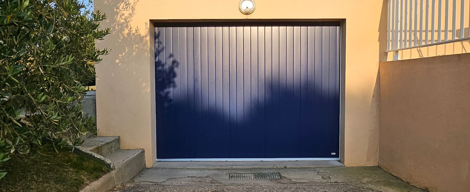 Une porte de garage bleu foncé avec des lignes verticales, des marches et un buisson.
