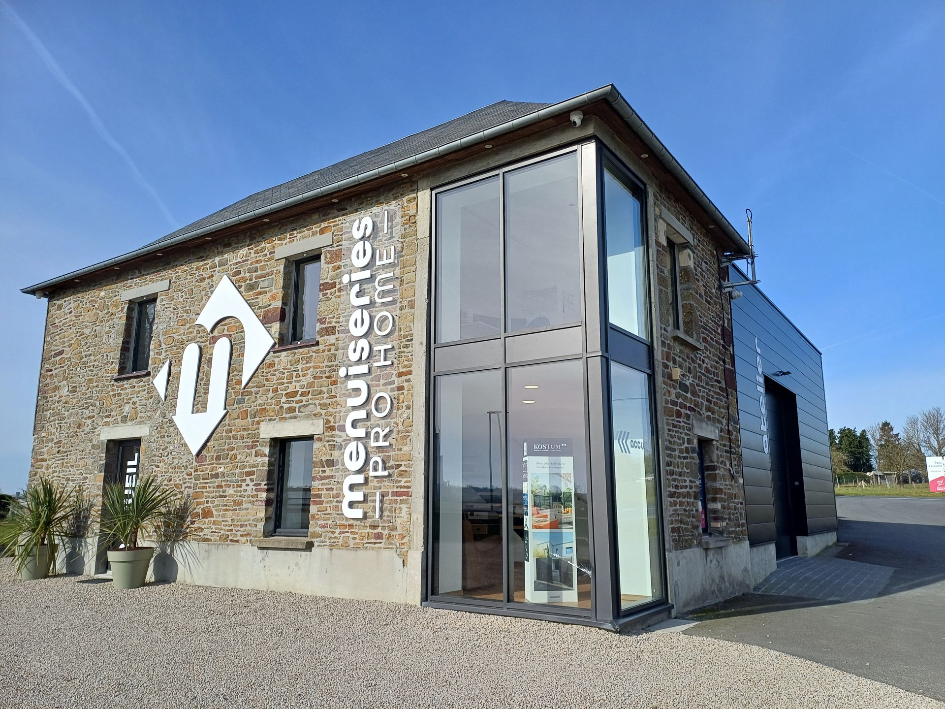 Bâtiment en pierre avec façade vitrée, enseigne « Menuiseries Pro Home ». Extérieur ensoleillé.