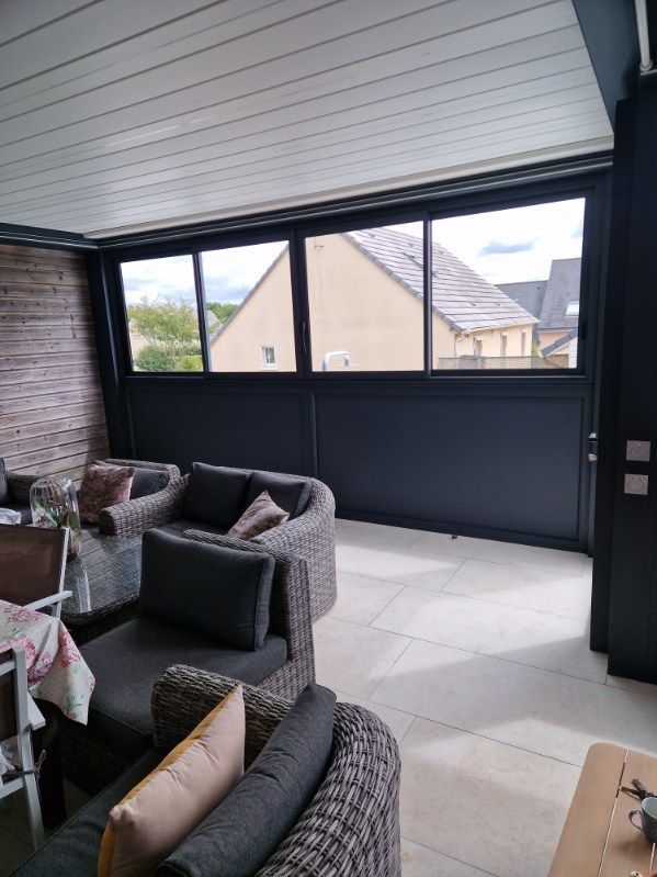 Espace salon intérieur avec mobilier gris foncé, grandes fenêtres donnant sur un bâtiment et un mur en pierre.