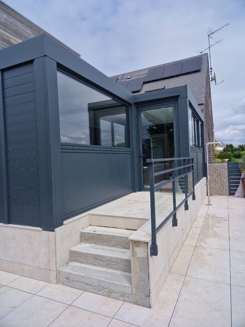 Véranda grise avec marches, panneaux de verre et balustrade en métal fixée à l'extérieur en pierre claire d'une maison.