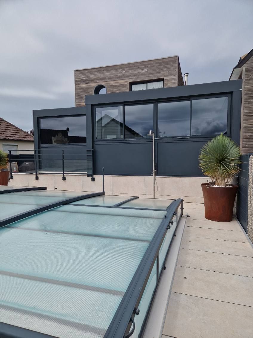 Terrasse sur le toit avec clôtures gris foncé, baies vitrées et piscine couverte. Ciel nuageux.