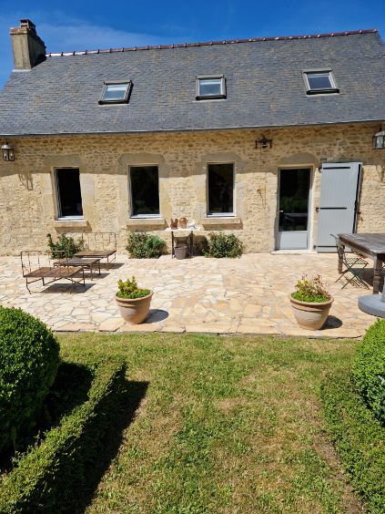Maison en pierre avec patio, jardin et trois lucarnes sous un ciel bleu.