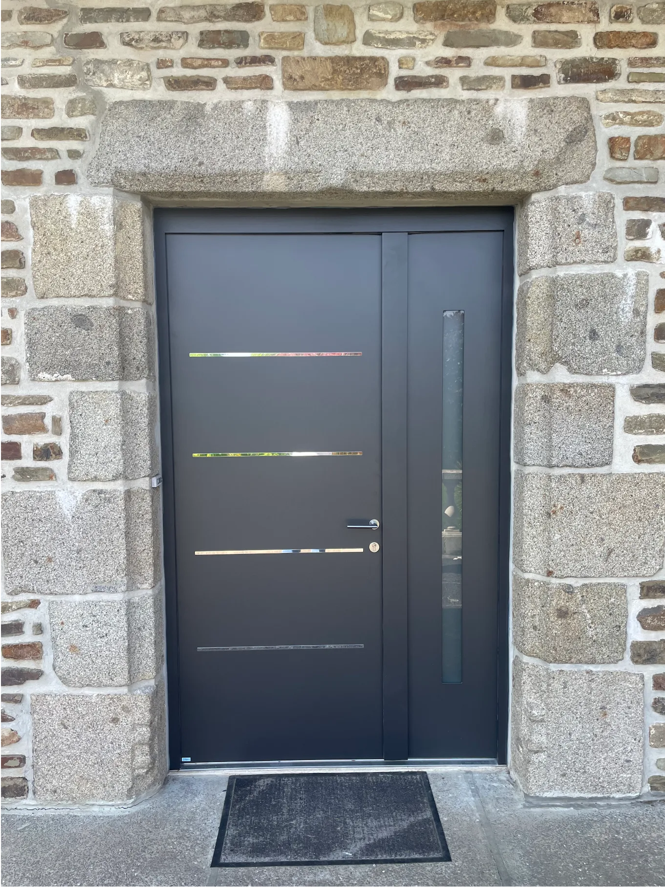 Porte gris foncé avec panneau latéral encastré dans un cadre en pierre, petit paillasson.