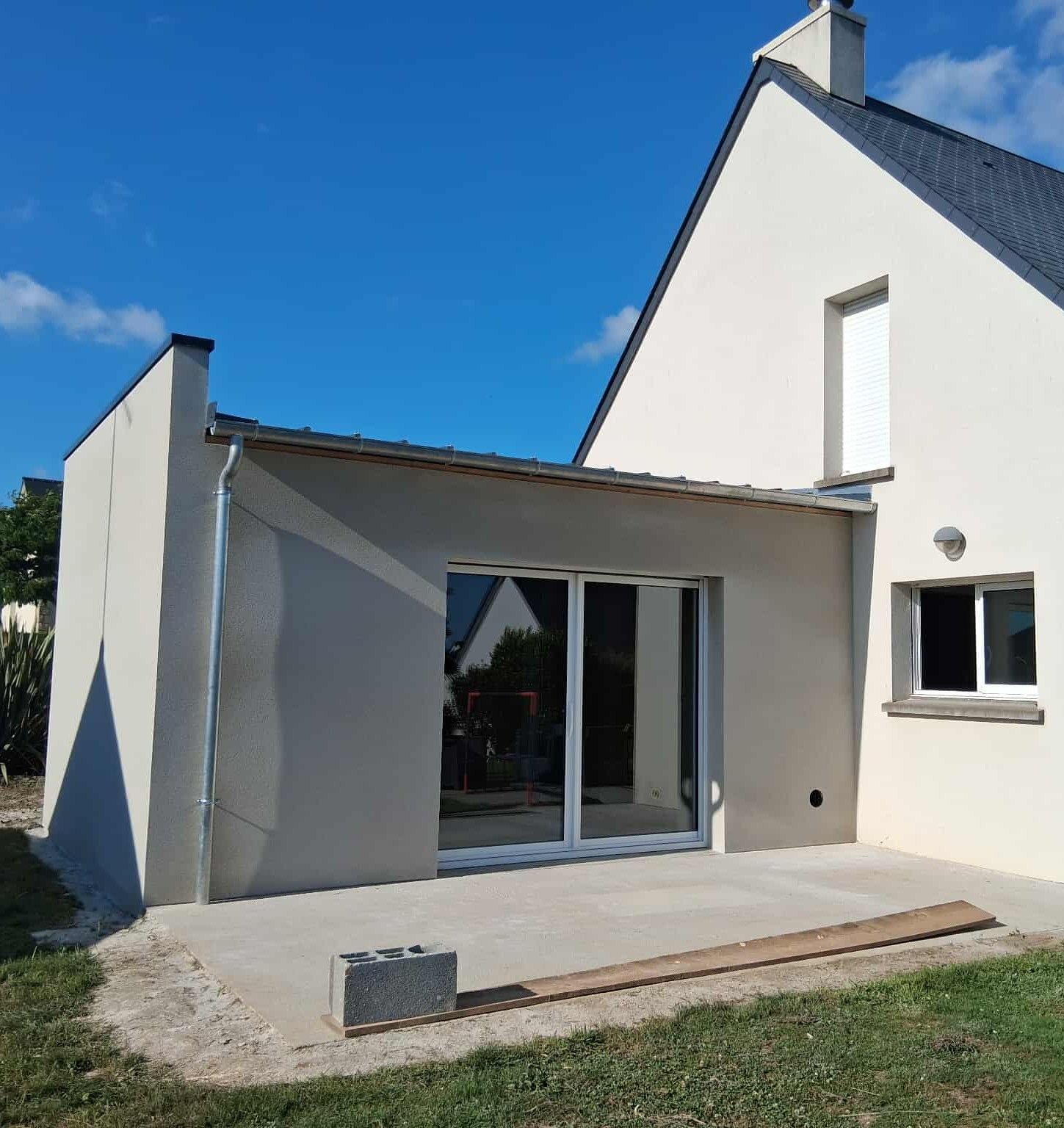 Vue extérieure d'une maison aux murs gris clair avec une extension moderne, une porte coulissante en verre et un patio en béton.