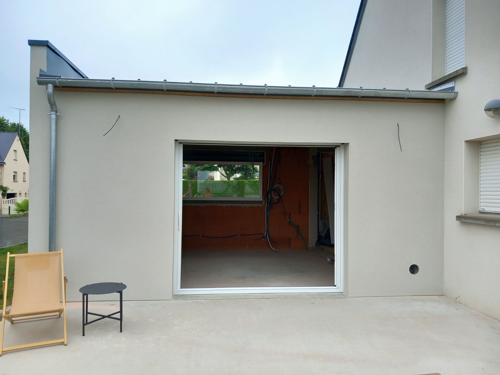 Extérieur d'une extension de maison avec une porte ouverte, des murs beiges et une terrasse en béton. Deux chaises sont installées à l'extérieur.