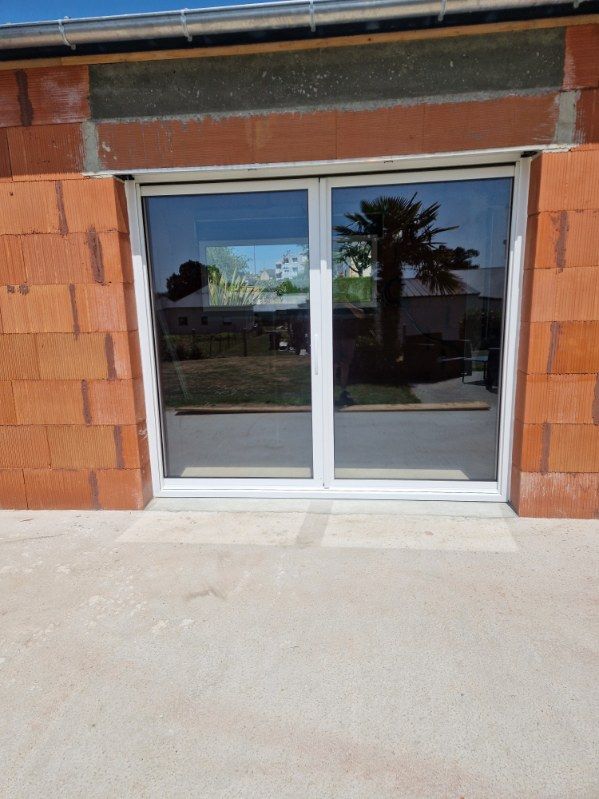 Porte coulissante en verre à cadre blanc dans un mur de briques en construction, avec arbre et ciel reflétés.