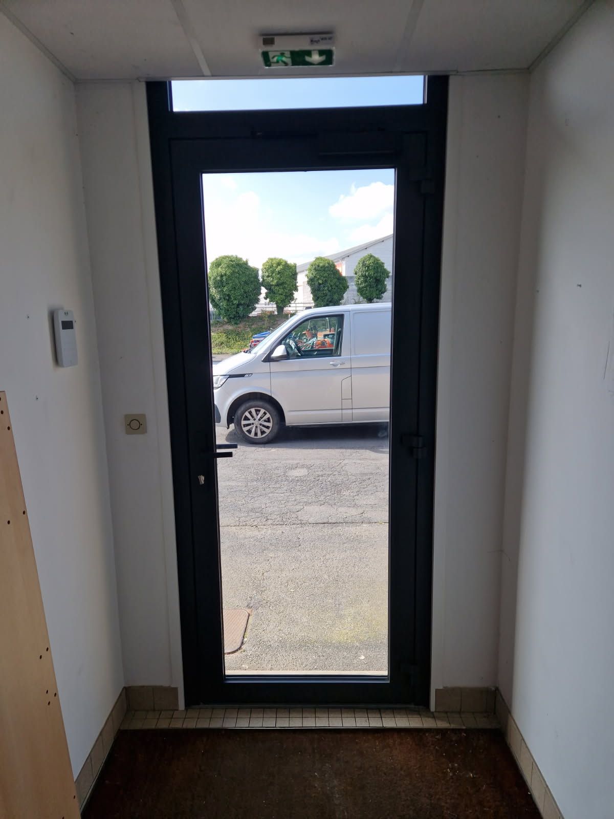 Porte vitrée à cadre noir ouverte sur l'extérieur avec une camionnette argentée garée. Panneau de sortie vert au-dessus.