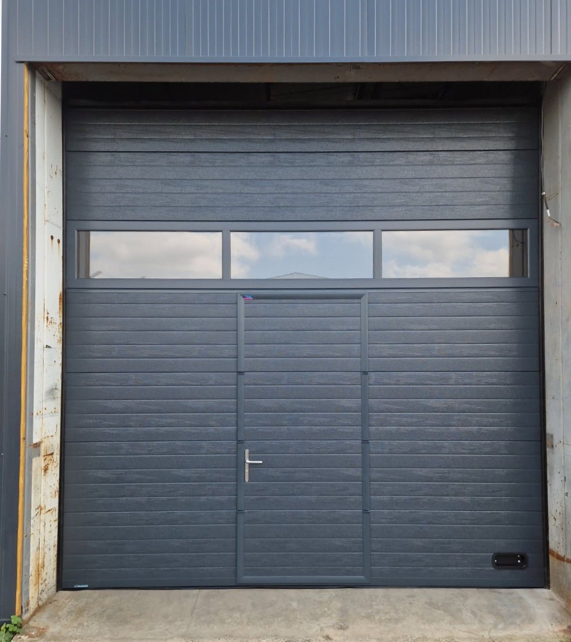 Porte de garage gris foncé avec une porte piétonne, trois fenêtres et une poignée argentée.