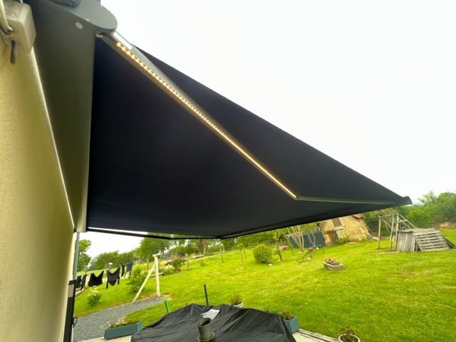 Auvent noir étendu depuis un mur, avec lumières intégrées, sur une cour herbeuse.