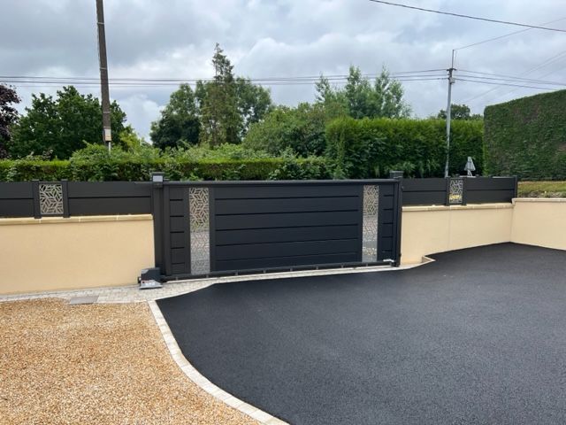 Portail coulissant noir avec inserts en pierre sur une allée pavée et mur beige.