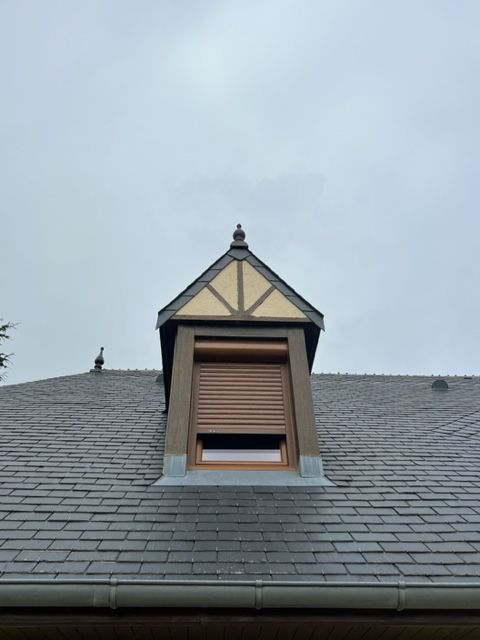 Lucarne à volet brun sur un toit en ardoise, sous un ciel gris.