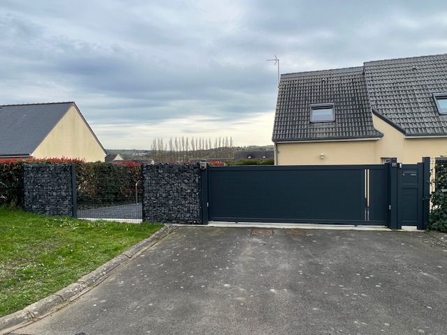 Un portail coulissant gris foncé et une clôture assortie avec des colonnes en pierre, devant les maisons.