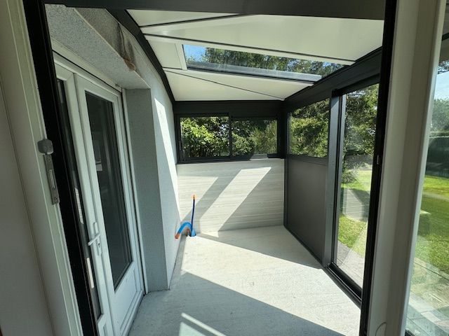 Véranda fermée avec toit en verre, porte ouverte et grandes fenêtres offrant une vue sur les arbres.