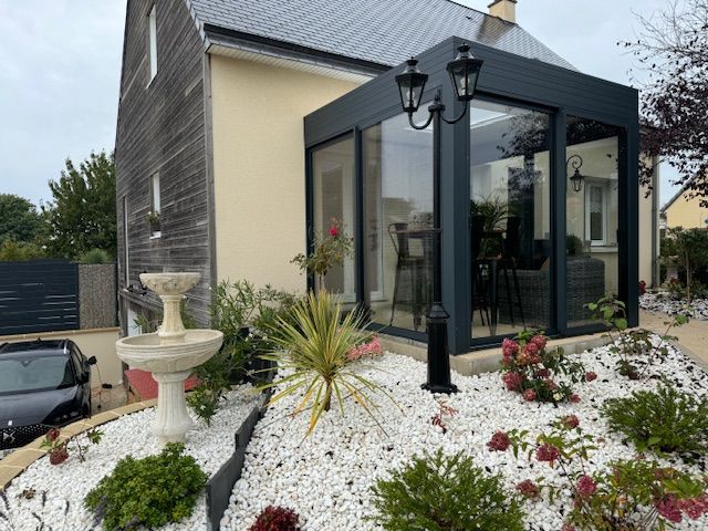 Une maison avec une véranda en verre à cadre sombre, dotée d'un jardin de rocaille blanc et d'une fontaine.