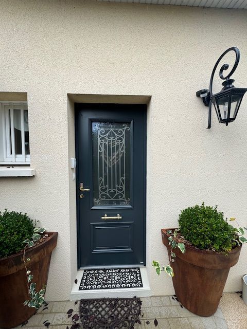 Porte d'entrée bleu foncé avec panneau de verre, flanquée de plantes en pot, placée dans un mur beige.