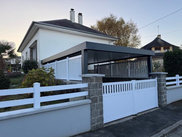 Maison blanche avec un abri voiture gris, une clôture blanche et un portail.