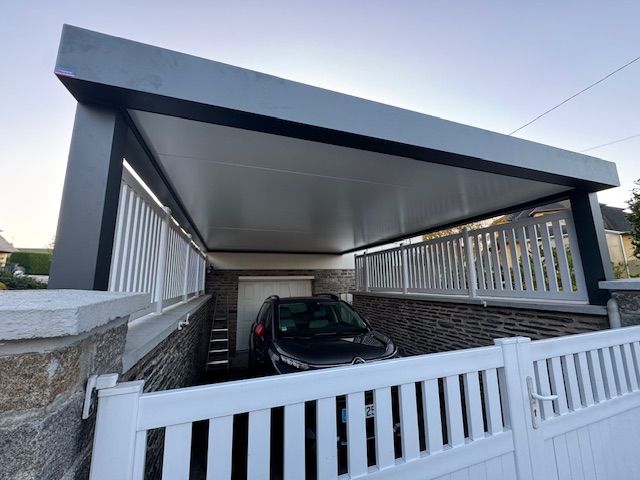 Carport avec une voiture garée en dessous, clôture blanche, toit et piliers gris, murs en pierre.