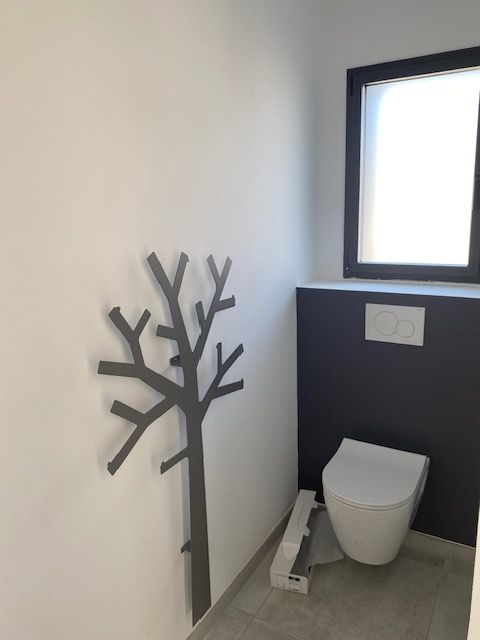 Salle de bain moderne avec murs blancs, toilettes, décoration murale en forme d'arbre gris foncé et fenêtre.